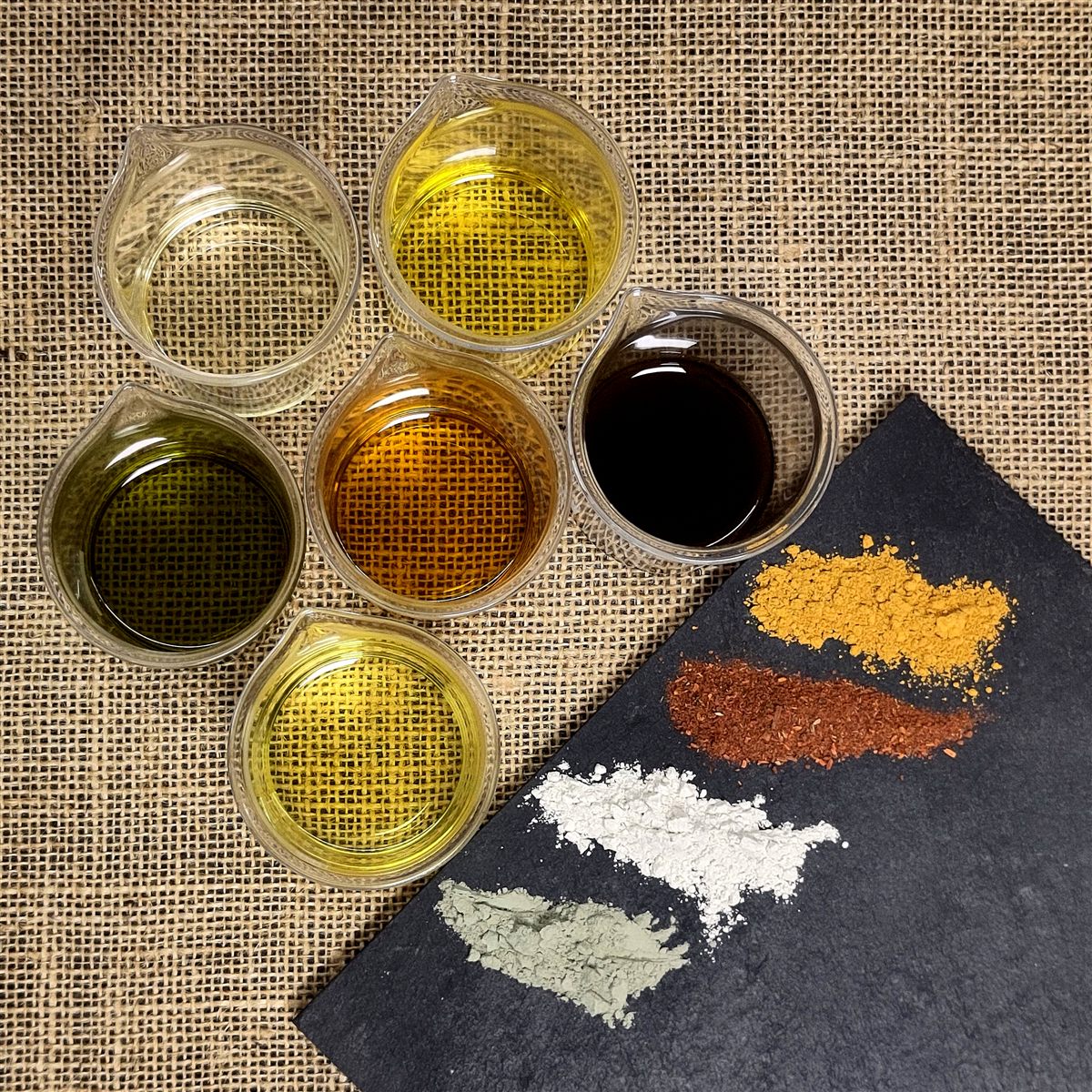 Six récipients en verre contenant différentes huiles végétales, accompagnés de poudres minérales colorées sur une toile de jute.